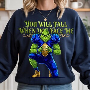 Peut inclure: Sweat-shirt bleu marine avec un personnage vert en tenue de football américain tenant un ballon. Le personnage est à genoux, avec le texte "YOU WILL FALL WHEN YOU FACE ME" en vert au-dessus. Le personnage porte des épaulettes et un pantalon bleus.
