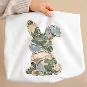 Op de afbeelding: Witte draagtas met een patchwork konijnontwerp in camouflagekleuren. Het konijn is gemaakt van stukken stof in groen, blauw en bruin. De tas wordt door een persoon vastgehouden. Het konijn heeft lange oren.