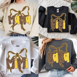 Könnte beinhalten: Vier Sweatshirts in verschiedenen Farben: Beige, Schwarz, Weiß und Grau. Jedes Sweatshirt zeigt ein Schleifendesign mit Leopardenmuster und die Wörter "Mama" oder "Mimi" in einem gelben, blasenartigen Schriftzug. Es handelt sich um Freizeitkleidung.