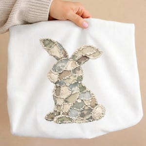 Op de afbeelding: Witte draagtas met een patchwork konijn ontwerp in groen, beige en blauw. Het konijn is geborduurd met een textuur, gestikt uiterlijk. De tas wordt door een persoon vastgehouden.