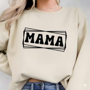 Op de afbeelding: Beige sweatshirt met het woord "MAMA" in vette, zwarte retro-letters in een rechthoekige lijst. De sweatshirt heeft lange mouwen en een ronde hals. Het ontwerp is gecentreerd op de voorkant van het kledingstuk.