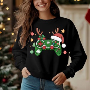 May include: Black sweatshirt featuring a green video game controller design with Christmas elements. The controller has reindeer antlers, a Santa hat, snowflakes, and gift box. Red, green, and white accents create a festive holiday theme.