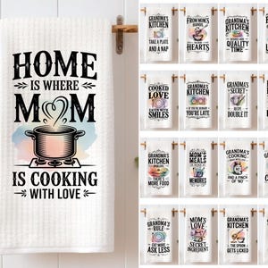 May include: White kitchen towel with the text "HOME IS WHERE MOM IS COOKING WITH LOVE" in black. The design includes a pot over a stove. Additional towels with kitchen-themed phrases are also displayed.