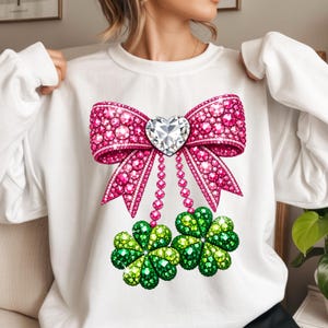 Könnte beinhalten: Weißes Sweatshirt mit einer großen, rosa verzierten Schleife mit herzförmiger Mitte. Unter der Schleife befinden sich zwei grüne, verzierte Kleeblätter. Das Design ist wahrscheinlich für St. Patrick's Day oder einen ähnlichen festlichen Anlass.