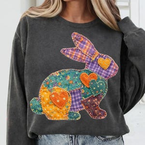 Op de afbeelding: Donkergrijze sweatshirt met een patchwork konijn ontwerp. Het konijn is samengesteld uit verschillende stoffen met patronen in tinten paars, teal, oranje en geel, met hart accenten. De sweatshirt heeft een ronde hals en lange mouwen.