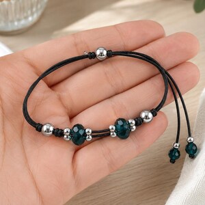 May include: A black cord bracelet featuring two faceted dark teal crystal beads, multiple small silver-toned metallic spheres, and two dangling ends each finished with a single dark teal crystal bead and a small silver-toned metallic sphere.