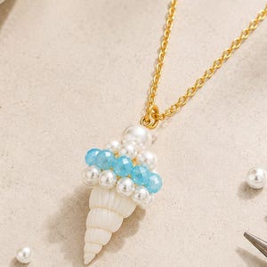 Op de afbeelding: Een goudkleurige ketting met een hanger in de vorm van een ijshoorntje, opgebouwd uit een witte spiraalvormige schelpbasis met daarboven afwisselende rijen kleine witte parels en gefacetteerde lichtblauwe kralen.