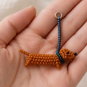 May include: A small, three-dimensional dachshund figurine crafted from tiny brown beads. The dog features a black bead for a nose and wears a dark blue beaded collar attached to a short, vertical blue beaded leash ending in a silver metal ring.