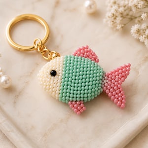 May include: A fish-shaped keychain crafted from small, round beads in cream, mint green, and pink. The fish features a single black bead for an eye and is attached to a polished gold-tone metal split ring by a short chain and a small gold bead.