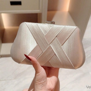 Puede incluir: Bolso de mano de satén beige con un diseño tejido en la parte delantera. El bolso rectangular tiene un cierre plateado y lo sostiene una mano con esmalte de uñas rojo. El bolso es de color neutro y parece estar hecho de un material suave.