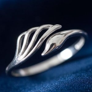 Può includere: Un anello d'argento dal design aperto, con un motivo stilizzato a foglia o piuma. L'anello è posto su un tessuto blu scuro, con perle e cristalli sparsi. Il design dell'anello è moderno e organico.