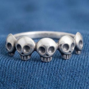 Può includere: Un anello d'argento con una serie di piccoli teschi. I teschi sono uniformemente distanziati attorno alla fascia, creando un motivo ripetuto. L'anello è posto su uno sfondo blu.