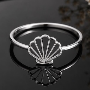 May include: A silver-toned ring featuring a delicate, open-work seashell motif centered on a thin, smooth band. The metal has a polished, reflective finish, and the design creates a clean, symmetrical silhouette of a fan-shaped scallop shell.