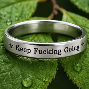 Op de afbeelding: Een zilverkleurige metalen ring met een gladde, gepolijste afwerking. De buitenkant van de band is gegraveerd met de tekst "Keep Fucking Going" in een duidelijk, zwart schreeflettertype, geflankeerd door een klein vijfpuntig sterretje aan weerszijden.