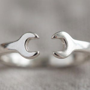 Puede incluir: Dos anillos plateados con forma de llave inglesa. Los anillos están hechos de un material metálico brillante y se colocan sobre un fondo suave y neutro. Las llaves se enfrentan, creando un diseño único. Estos anillos son un accesorio único.