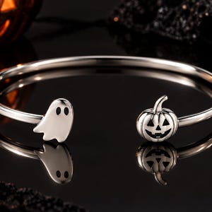 Op de afbeelding: Een zilverkleurige open armband met een kleine spookbedel aan het ene uiteinde en een uitgesneden pompoen-lantaarnbedel aan het andere uiteinde. Het metaal heeft een gepolijste afwerking en de bedels bevatten zwarte details voor de ogen.