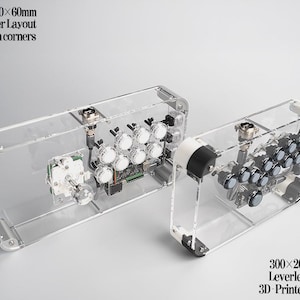 Könnte beinhalten: Zwei transparente Kampf-Sticks aus Acryl. Der größere ist 380 x 200 x 60 mm mit Hebel-Layout und Aluminiumecken. Der kleinere ist 300 x 200 x 60 mm mit leverless Layout und 3D-gedruckten Ecken.