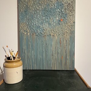 Puede incluir: Pintura abstracta texturizada en tonos azules, grises y naranjas. La obra de arte presenta un patrón de formas circulares en relieve en la parte superior, que se transforman en pinceladas verticales. Un tarro de cerámica con pinceles está a la izquierda.