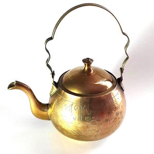 May include: A vintage brass teapot with a curved spout and a decorative handle. The teapot features etched floral patterns and a lid with a small knob. The overall colour is a warm gold.