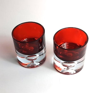 May include: Two cylindrical drinking glasses with a red interior and a clear, rounded base. The glasses are made of glass and are set against a white background. The glasses are empty.