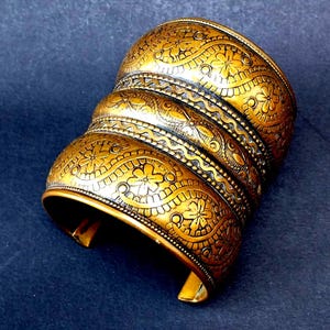 May include: A wide gold-toned metal cuff bracelet featuring three distinct horizontal bands. Each band is decorated with an embossed pattern of small flowers and swirling vine motifs. The bracelet has an open back design with a polished finish.