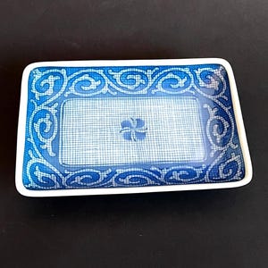 May include: Rectangular blue and white dish with a floral design in the centre. The dish has a white border and a decorative blue pattern around the edge. The background is a dark surface.