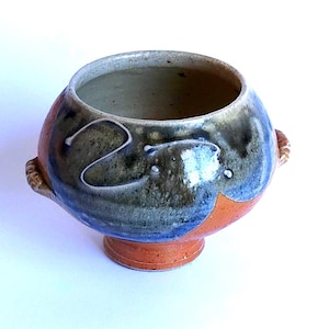 May include: A handmade ceramic bowl with a rounded form and a small, circular base. The bowl features a colour palette of blue, brown, and orange, with a white, abstract design. It has two small, textured handles on either side.