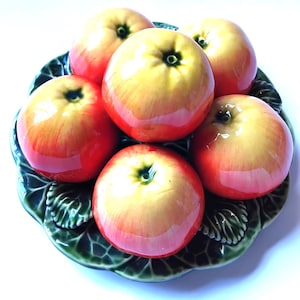 Peut inclure: Une assiette décorative en céramique en forme de grande feuille verte, surmontée de cinq pommes rondes, brillantes et réalistes. Les pommes présentent un dégradé de jaune et de rouge, tandis que l'assiette a un motif de feuille texturé vert foncé.