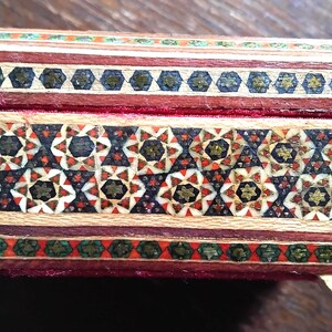 May include: A rectangular wooden box featuring intricate geometric marquetry patterns in shades of red, black, white, and green. The lid and body are decorated with star-shaped motifs and linear borders, with a visible red velvet interior lining.
