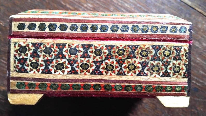 May include: A rectangular wooden box featuring intricate geometric marquetry patterns in shades of red, black, white, and green. The lid and body are decorated with star-shaped motifs and linear borders, with a visible red velvet interior lining.
