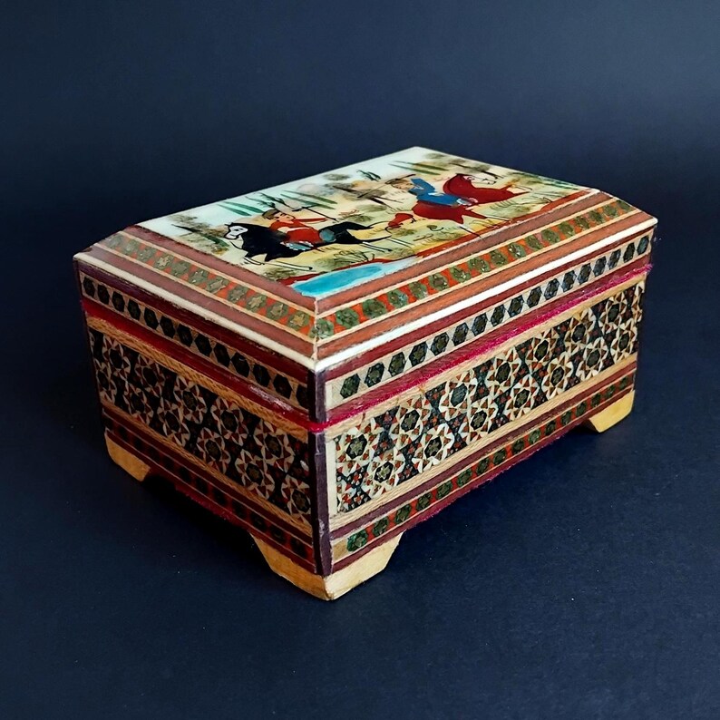 May include: A rectangular wooden box featuring intricate geometric marquetry patterns on the sides and a painted lid depicting figures on horseback in a landscape. The box is supported by four small feet and displays a multi-colored inlay design.