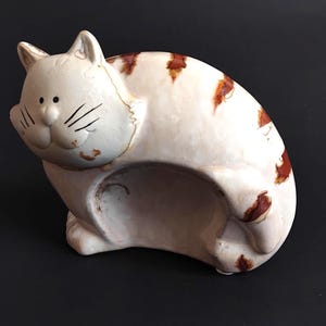 Può includere: Statuetta di gatto in ceramica in posizione seduta, con base bianca e segni marroni. Il gatto ha una faccia sorridente con baffi neri e riposa sulle zampe posteriori. La statuetta ha un aspetto invecchiato.