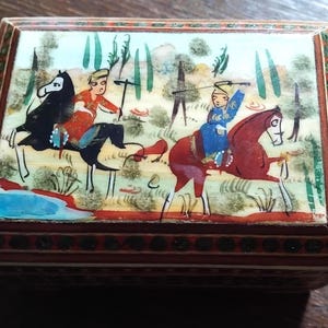 May include: A rectangular wooden box featuring a hand-painted lid depicting two figures on horseback in a landscape with trees and a blue pond. The box has a dark brown frame with intricate geometric patterns and a decorative border around the lid.