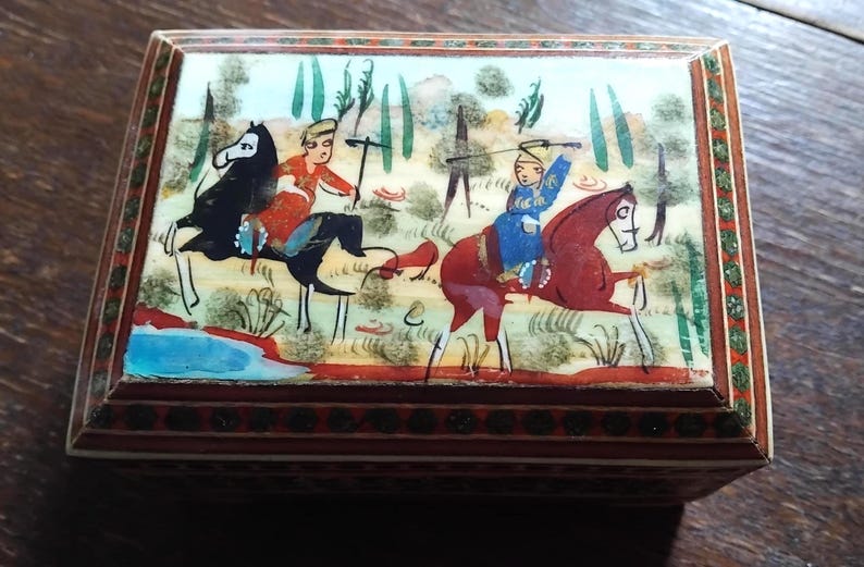 May include: A rectangular wooden box featuring a hand-painted lid depicting two figures on horseback in a landscape with trees and a blue pond. The box has a dark brown frame with intricate geometric patterns and a decorative border around the lid.