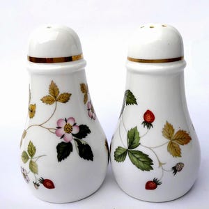 Peut inclure: Une paire de salière et poivrière en porcelaine blanche de forme conique. Chaque pièce présente un liseré doré autour du col et un motif botanique composé de feuilles vertes, de petites fleurs roses et de fraises rouges sur fond blanc.