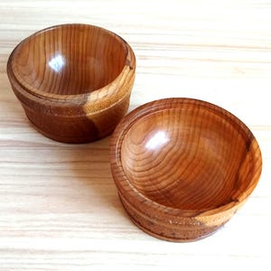 Puede incluir: Dos cuencos de madera hechos a mano con un acabado de veta natural. Los cuencos son redondos con un borde ligeramente ensanchado y un interior liso. La madera tiene un tono marrón rojizo cálido. Los cuencos parecen de tamaño mediano.