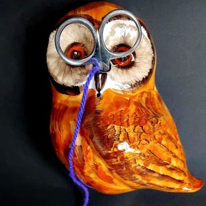 May include: A ceramic owl figurine with orange and brown hues, holding a pair of silver scissors and a strand of purple yarn. The owl has large, expressive eyes and is set against a dark background. A unique decorative item for a craft room.