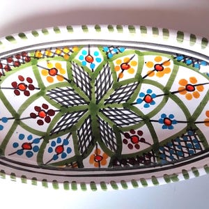 Pode incluir: Travessa oval de cerâmica com base branca e design floral colorido. O design inclui flores verdes, azuis, laranjas e vermelhas, com um centro geométrico verde e preto. A borda é branca com uma borda pontilhada verde.