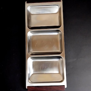Puede incluir: Una bandeja de servir rectangular con tres compartimentos separados de acero inoxidable dispuestos verticalmente. La bandeja está enmarcada por asas de madera de color marrón oscuro en los extremos superior e inferior, contrastando con el metal.