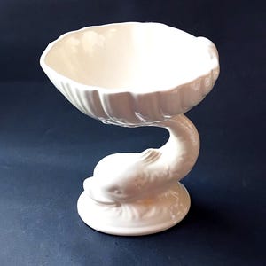 May include: A glossy off-white ceramic pedestal bowl featuring a sculpted dolphin base. The bowl has a scalloped, shell-like rim and rests on a circular foundation. The entire piece is finished in a smooth, reflective glaze with detailed moulding.