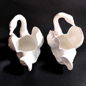 Puede incluir: Un par de maceteros o recipientes de cerámica blanca con forma de cisne. Cada pieza presenta un cisne esculpido con un cuello largo y curvado y texturas de alas detalladas que forman el contenedor abierto. El acabado blanco brillante resalta los patrones.