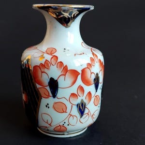 May include: Small white ceramic vase with orange floral design and blue accents. The vase has a narrow neck with gold trim and a flared rim. The design includes orange flowers, blue leaves, and gold accents. The vase is likely for decorative purposes.
