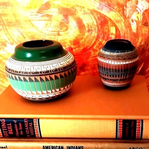 Op de afbeelding: Twee keramische potten met geometrische patronen. De grotere pot is groen, wit en bruin met een brede opening. De kleinere pot is bruin, zwart en wit met een smalle opening. Ze staan op een boek met de titel Sitting Bull Champion of the Sioux.