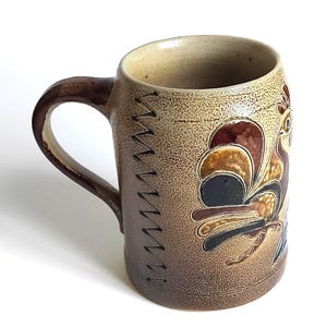 May include: A ceramic mug with a brown handle and a textured, speckled surface. The mug features a rooster design in shades of brown, black, and blue. Decorative stitching accents the side of the mug.