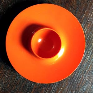 May include: Bright orange egg cup with a wide, flat base and a central, rounded cup. The egg cup is made of a glossy material and sits on a dark wooden surface. The design is simple and modern.