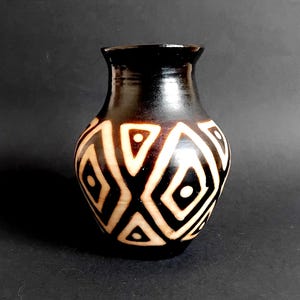 May include: A small, rounded ceramic vase with a glossy black finish. The body of the vase is decorated with a repeating pattern of cream-colored diamonds and triangles, each featuring a central dot, creating a high-contrast geometric design.