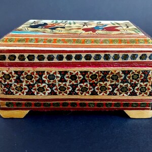 May include: A rectangular wooden box featuring intricate marquetry with geometric star patterns in red, black, and cream. The lid displays a painted scene of figures on horseback. The box has a hinged top and rests on four small, light-colored wooden feet.
