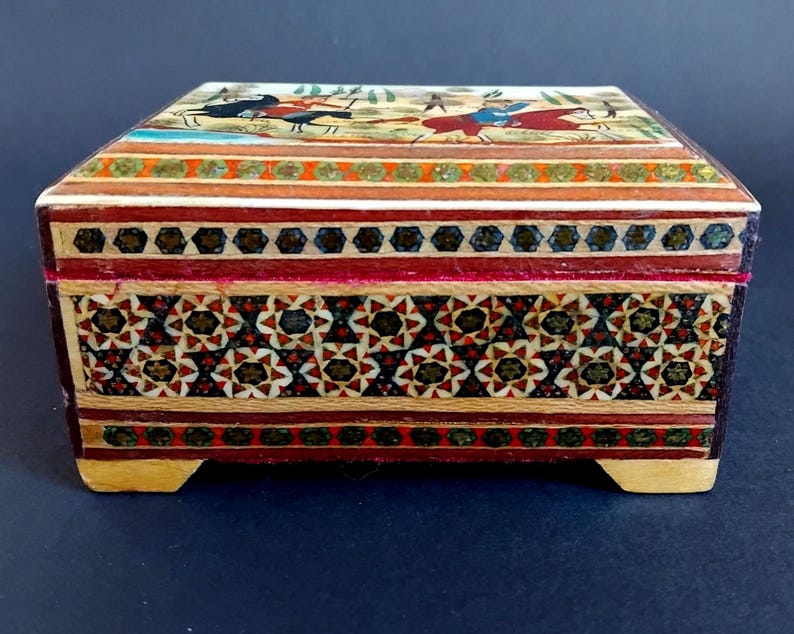 May include: A rectangular wooden box featuring intricate marquetry with geometric star patterns in red, black, and cream. The lid displays a painted scene of figures on horseback. The box has a hinged top and rests on four small, light-colored wooden feet.
