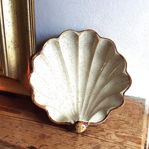 May include: A ceramic dish shaped like a seashell with a cream-coloured glaze and dark brown speckles throughout. The dish features deep, radiating ridges and a smooth, glossy finish, with a darker brown accent at the base of the shell structure.
