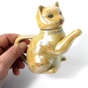 Puede incluir: Una tetera de gato vintage caprichosa en un tono amarillo claro. La tetera presenta una cabeza de gato como tapa y está adornada con pequeños patrones florales en rojo, azul y verde. Tiene un asa y un pico diseñado como un brazo de gato.